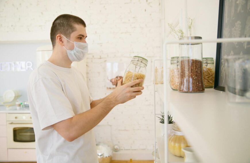 A young man with a facemask in a modern kitchen reviewing pantry items.