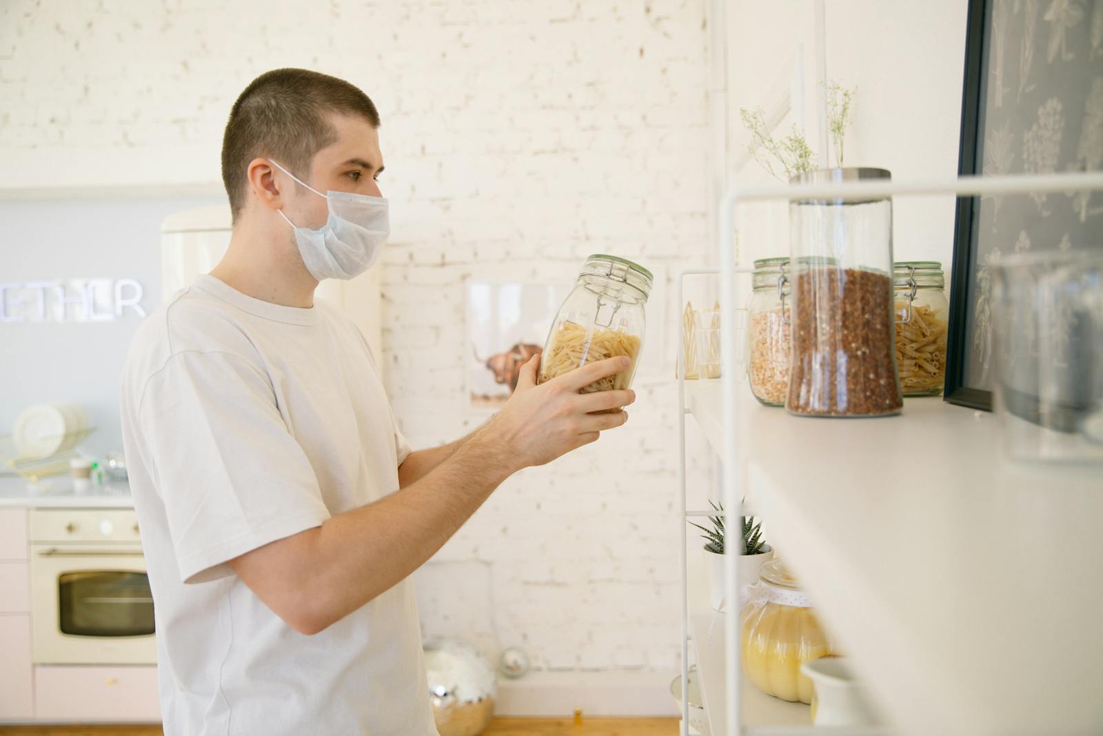 A young man with a facemask in a modern kitchen reviewing pantry items.