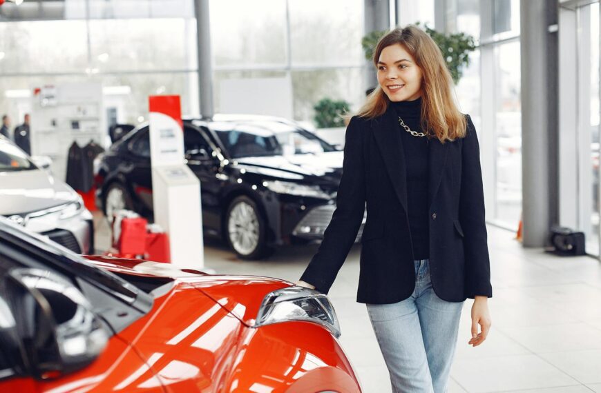 Happy stylish young female in jeans and black jacket standing near modern red shiny car in contemporary car showroom and looking at purchase dreamily