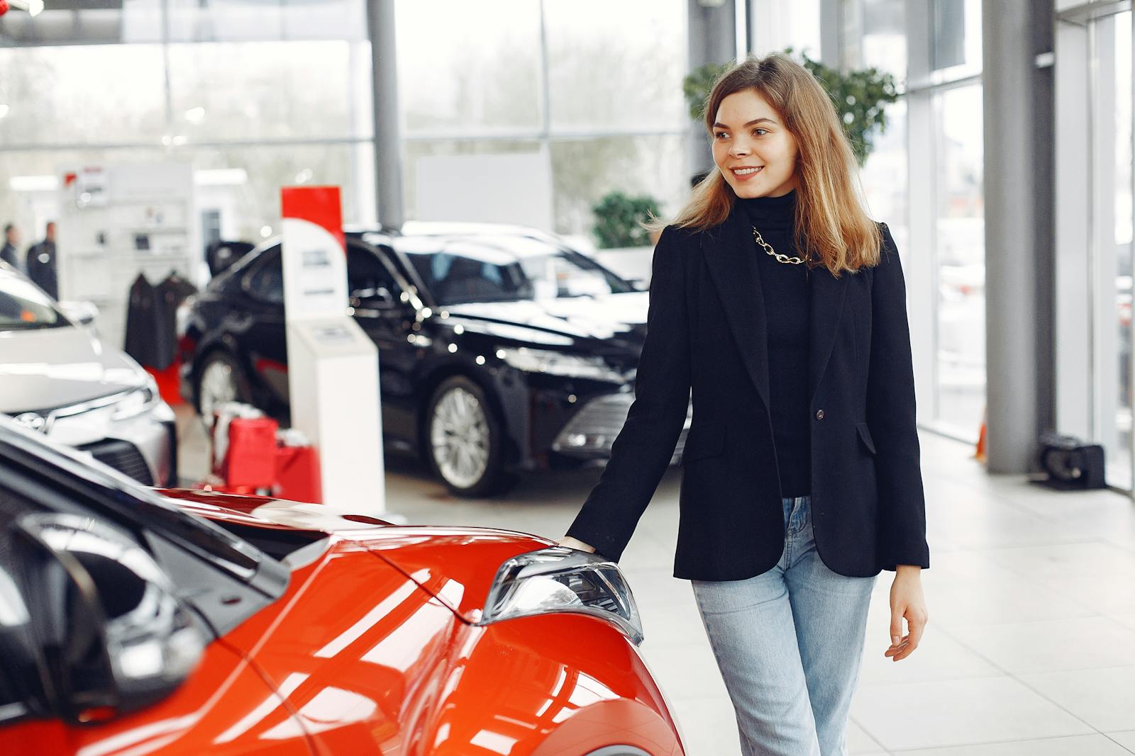 Happy stylish young female in jeans and black jacket standing near modern red shiny car in contemporary car showroom and looking at purchase dreamily