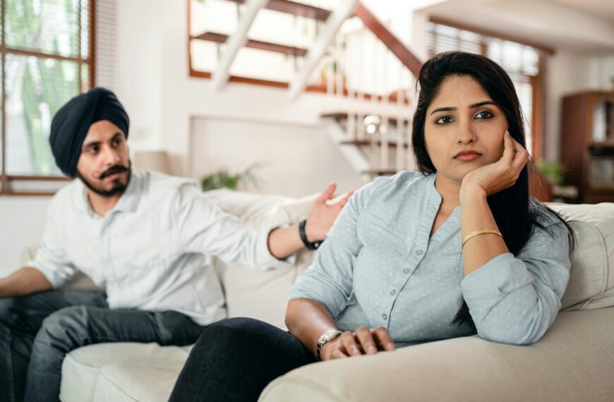 Young bearded Sikh man in casual wear and turban arguing with sad wife sitting on sofa