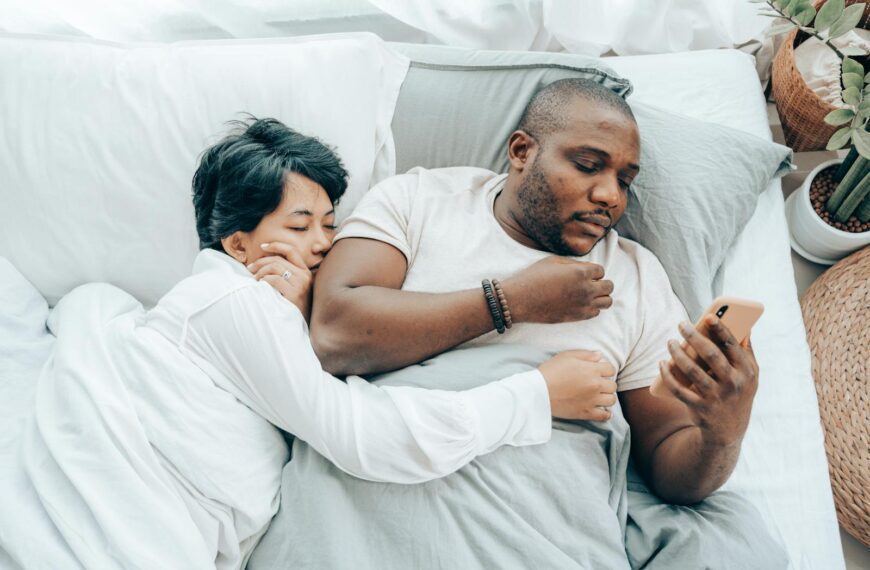 From above of young sleeping Asian woman cuddling black husband reading message on smartphone
