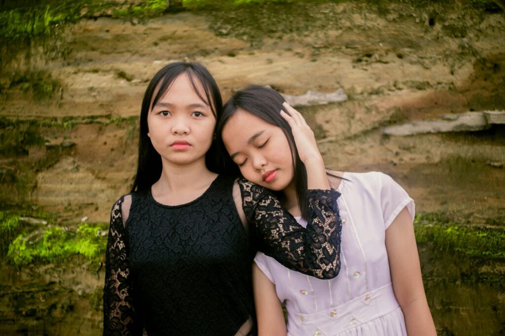 Calm Asian teen girl in black dress hugging girlfriend while standing close to each other