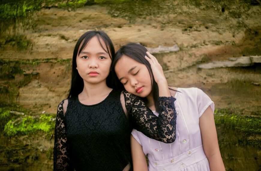 Calm Asian teen girl in black dress hugging girlfriend while standing close to each other
