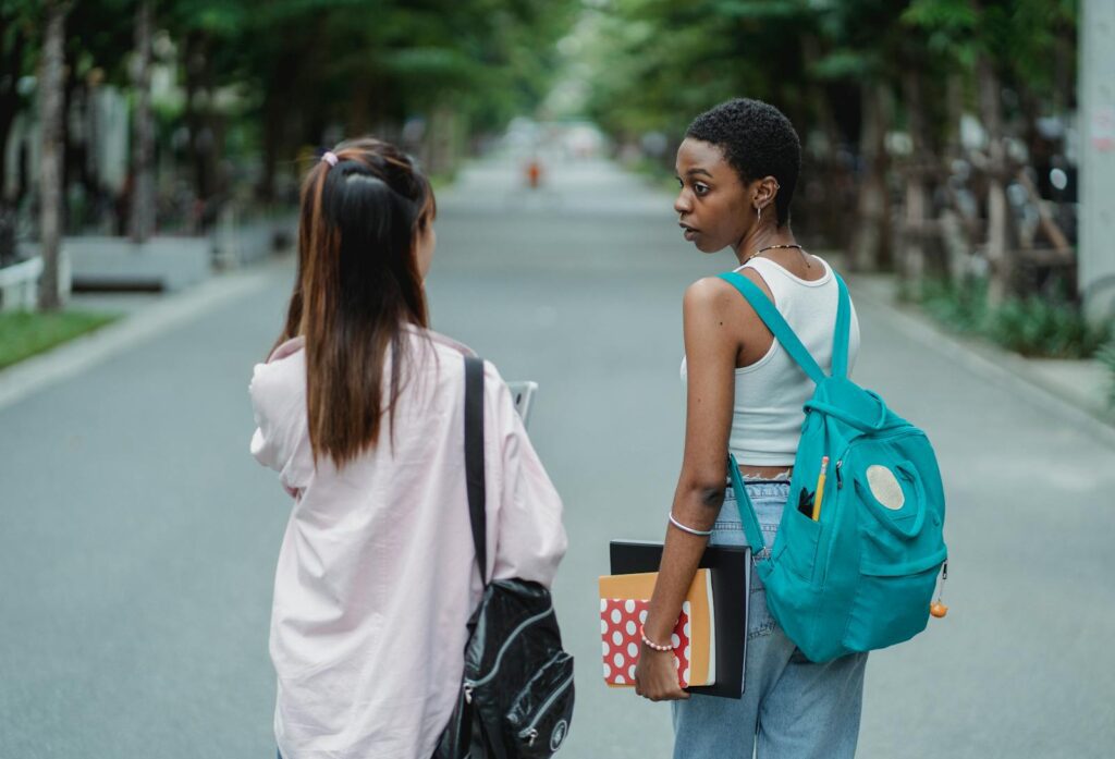 Back view of diverse female students carrying backpacks and supplies for university while talking in park