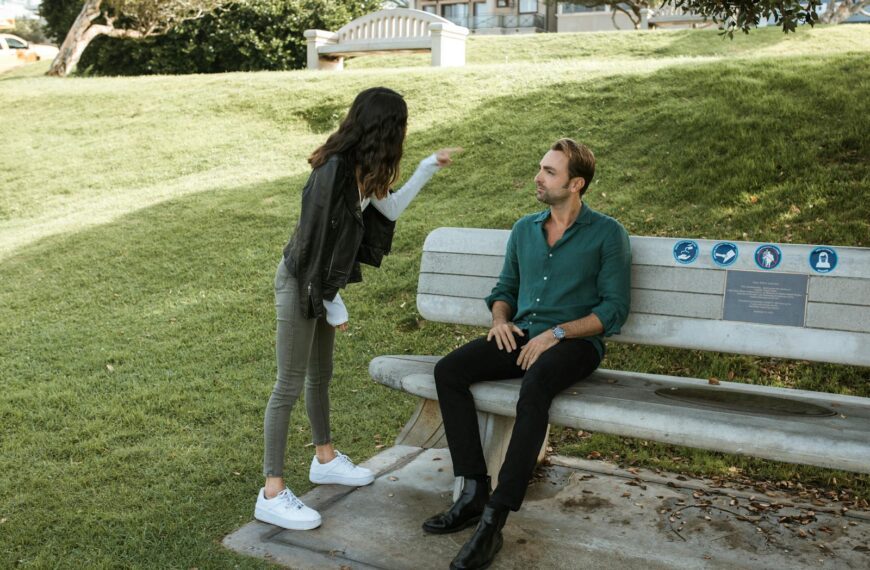 A couple in a heated argument while sitting and standing in a sunny park setting.
