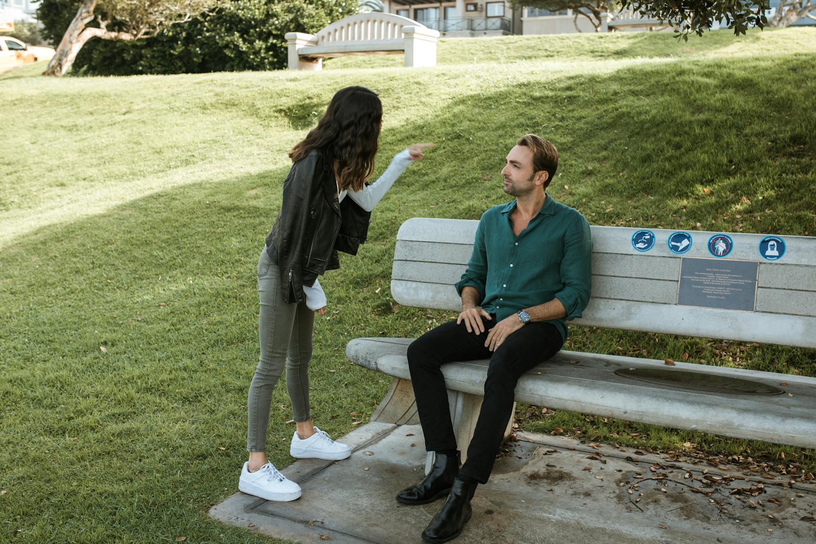 A couple in a heated argument while sitting and standing in a sunny park setting.