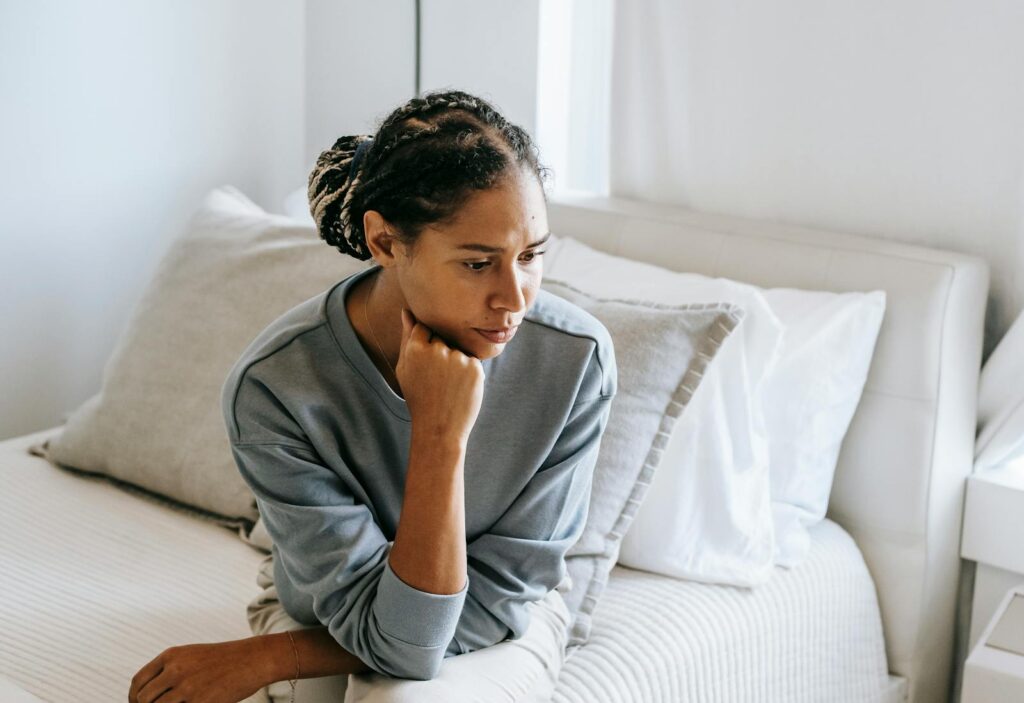 Serious young African American lady in casual clothes sitting on white bed and thinking while looking down in bedroom