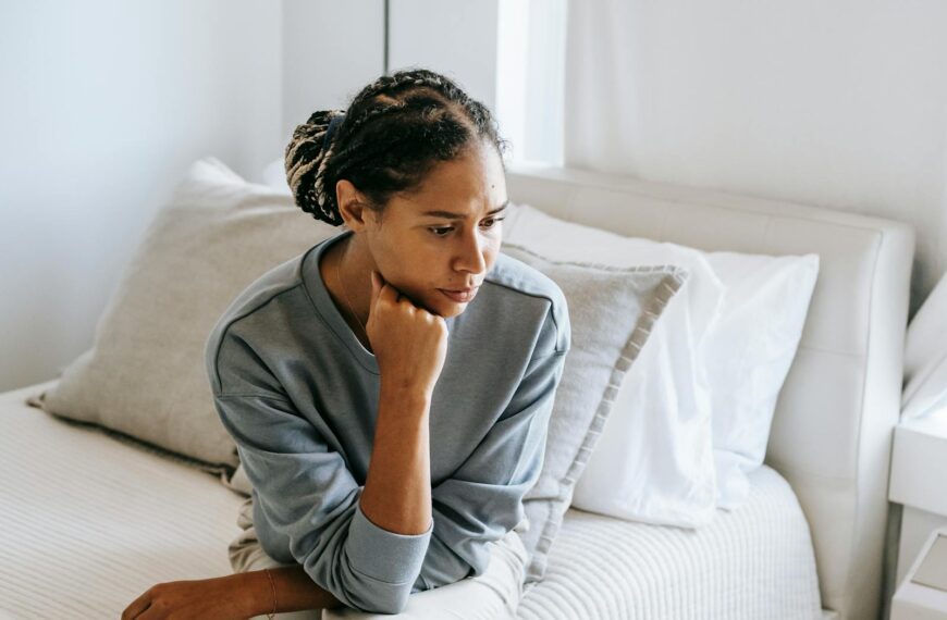 Serious young African American lady in casual clothes sitting on white bed and thinking while looking down in bedroom