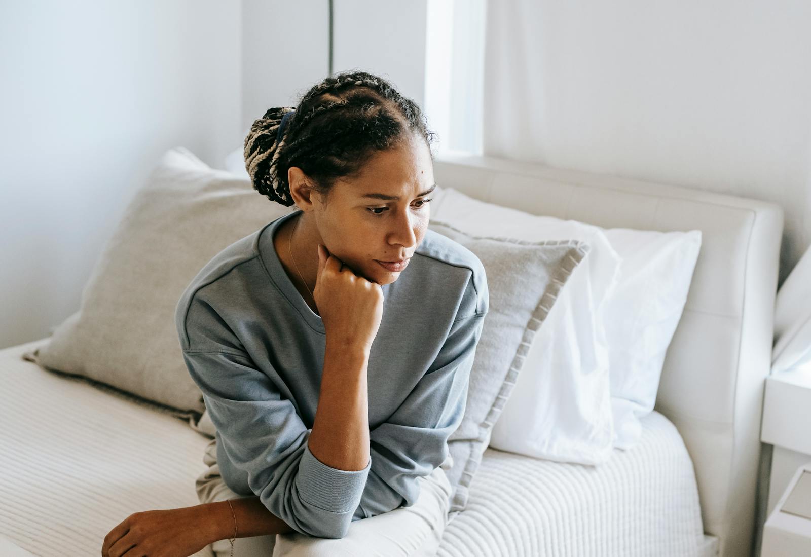 Serious young African American lady in casual clothes sitting on white bed and thinking while looking down in bedroom