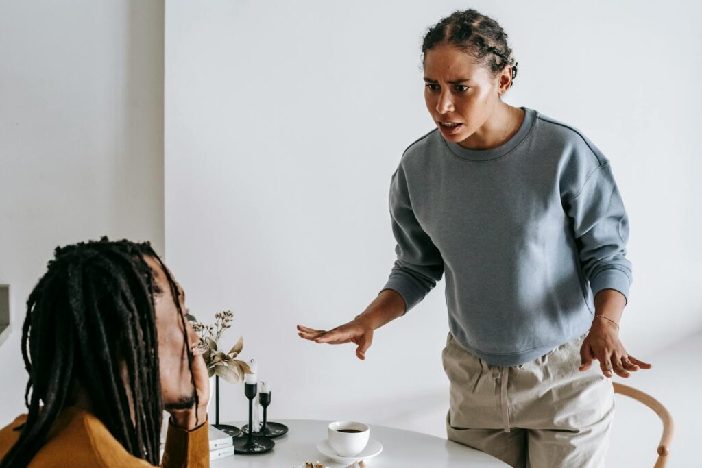 Serious young African American female in casual outfit standing near round table with cup of coffee and arguing with bearded husband