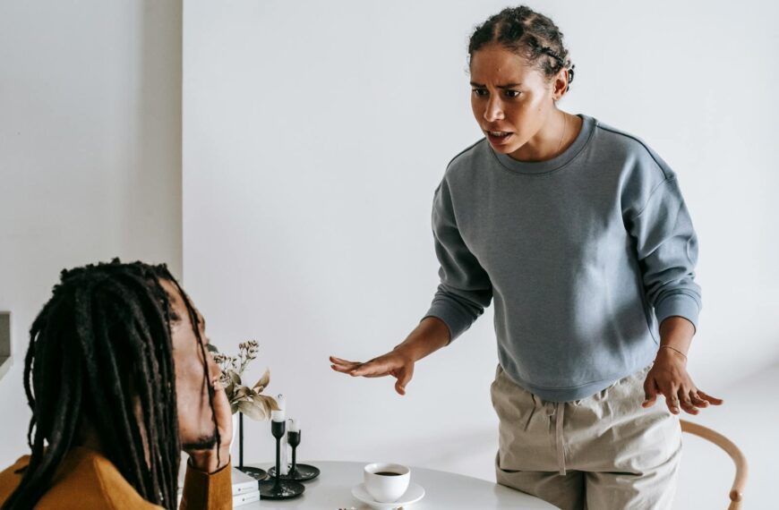 Serious young African American female in casual outfit standing near round table with cup of coffee and arguing with bearded husband