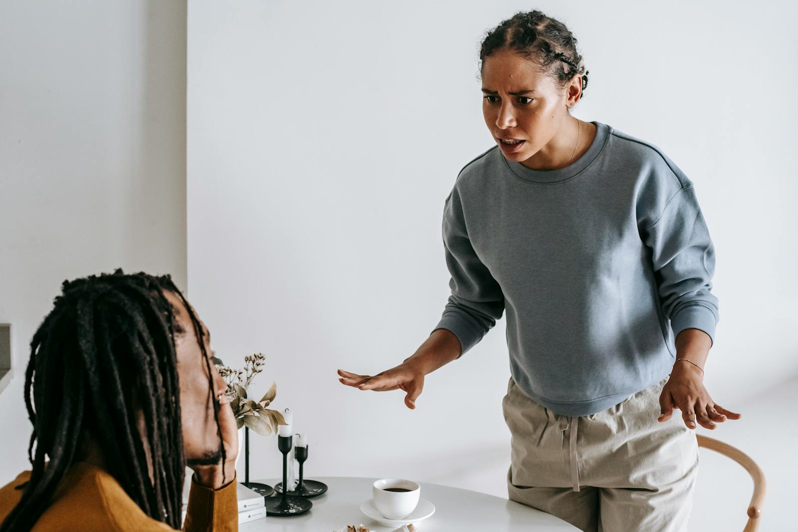 Serious young African American female in casual outfit standing near round table with cup of coffee and arguing with bearded husband