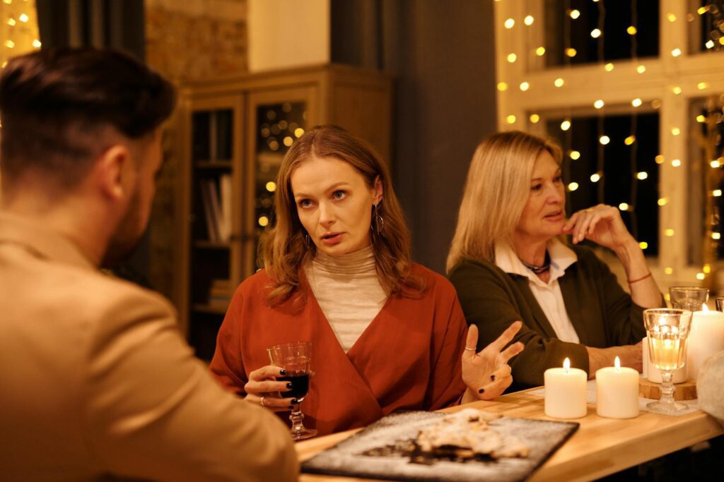 Three people enjoying a festive dinner table conversation surrounded by warm holiday lights.