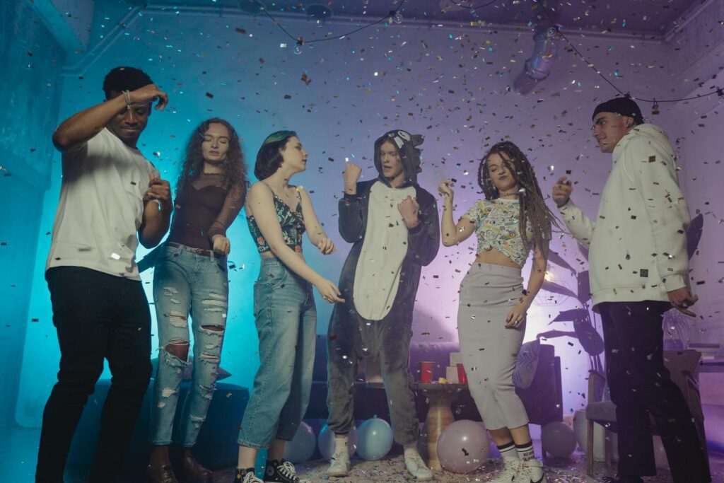 A group of diverse young adults dancing and having fun at an indoor party with confetti and balloons.