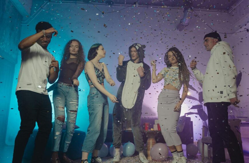 A group of diverse young adults dancing and having fun at an indoor party with confetti and balloons.