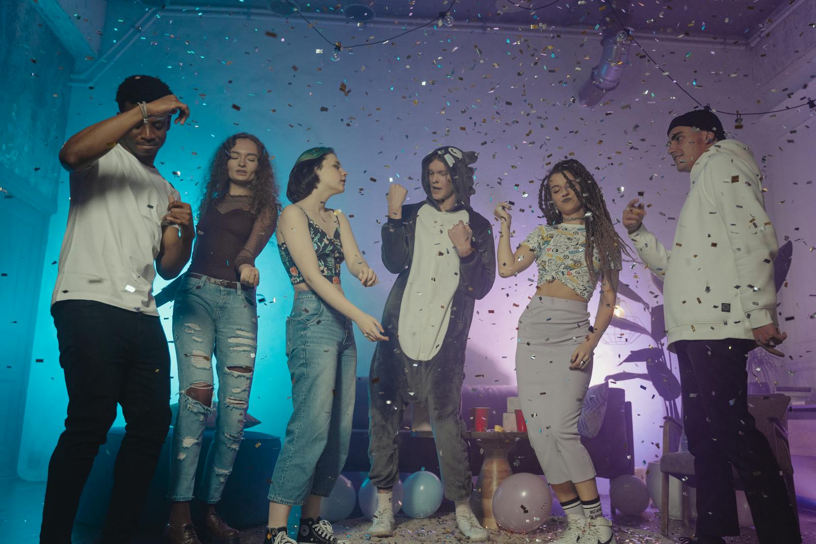 A group of diverse young adults dancing and having fun at an indoor party with confetti and balloons.