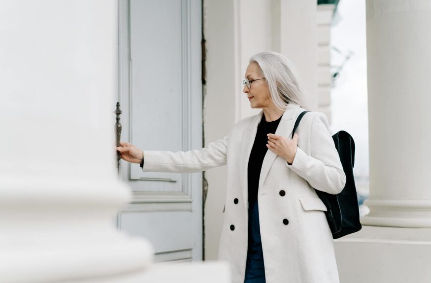 Stylish senior woman in white coat opening a door while standing outdoors, exuding confidence and grace.