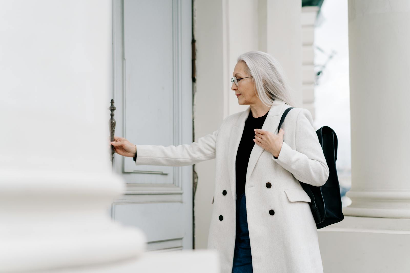 Stylish senior woman in white coat opening a door while standing outdoors, exuding confidence and grace.
