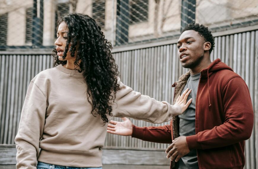 A couple in casual clothing appears to be in a disagreement outdoors with grey fencing background.