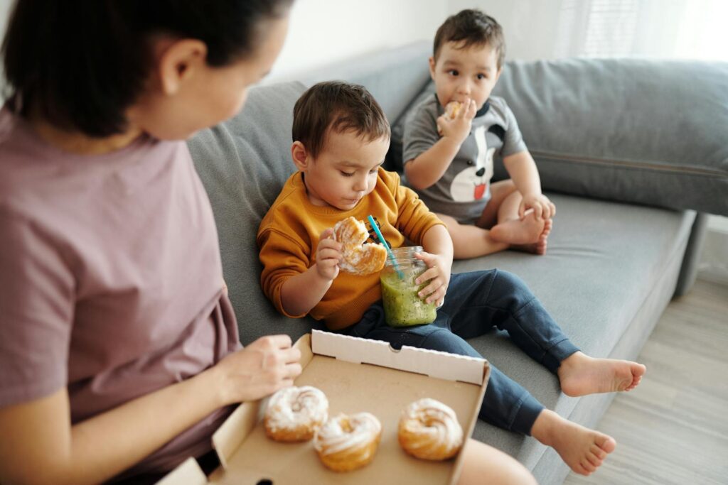 Mother and sons sitting on sofa, enjoying pastries and smoothies indoors.