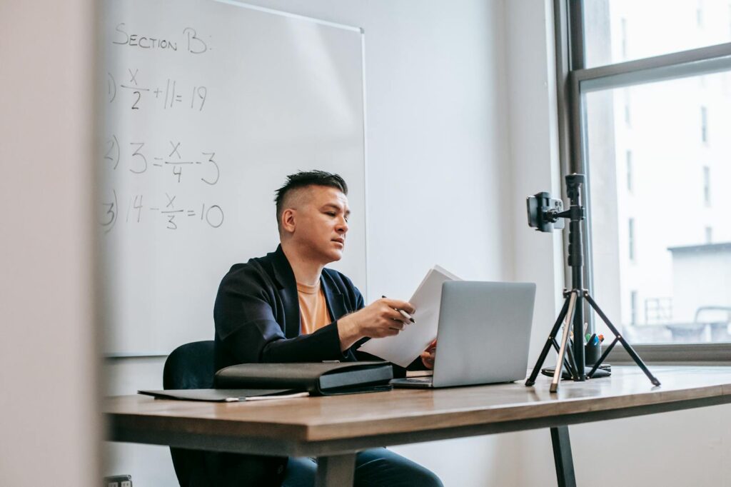 A male teacher conducting an online class with a laptop and camera setup in a classroom.