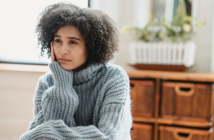 Discontent African American female leaning on hand and looking away while sitting in light room near green plant on blurred background