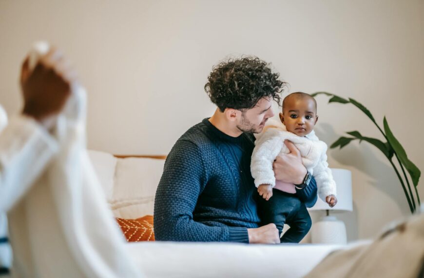 Content young man with curly hair in stylish sweater hugging adorable ethnic baby while resting on sofa near crop unrecognizable wife