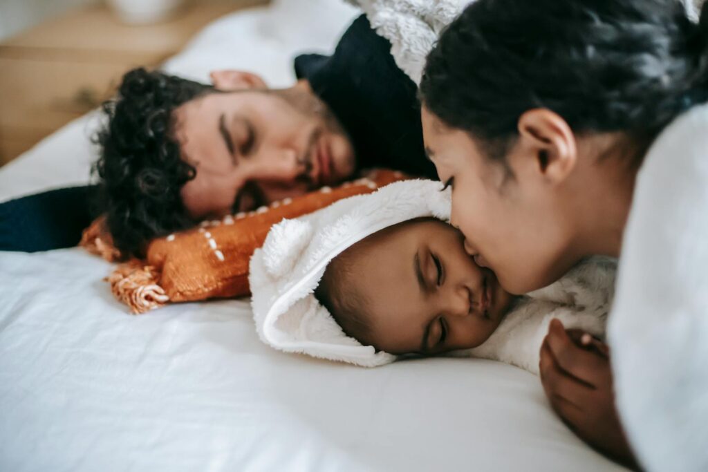 From above of crop ethnic lady with dark hair kissing cheek of sweet newborn baby lying in bed near sleeping father at home