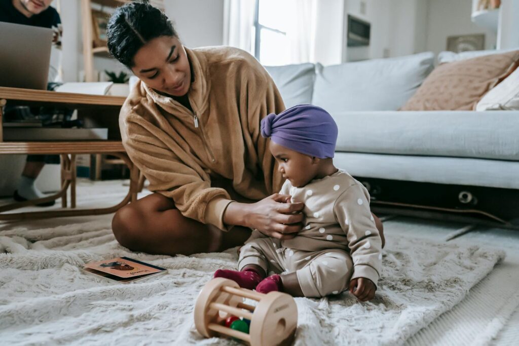 Full body of content African American mother and little baby sitting on floor with toys while spending time in living room with couch