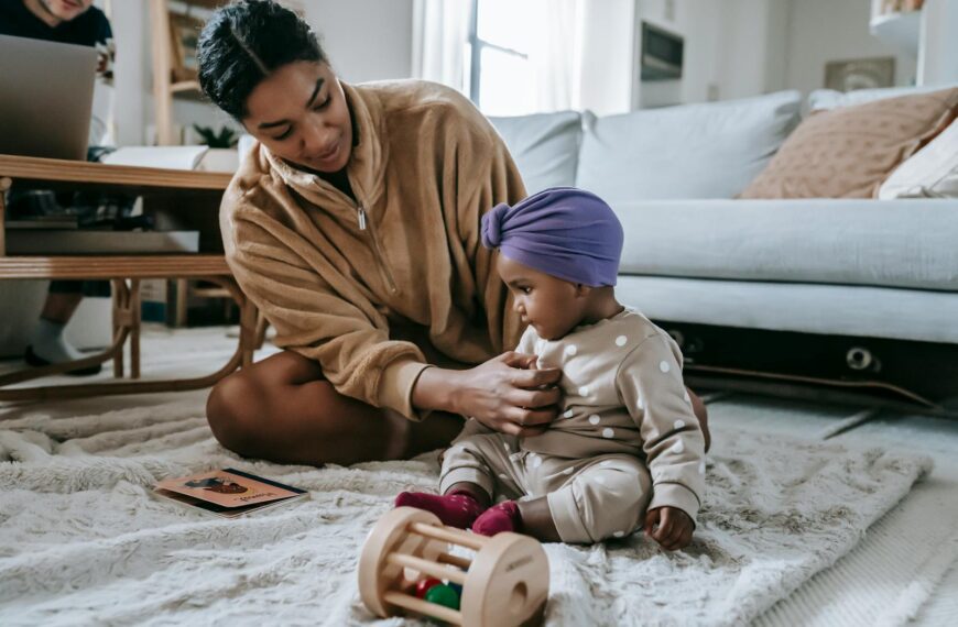 Full body of content African American mother and little baby sitting on floor with toys while spending time in living room with couch
