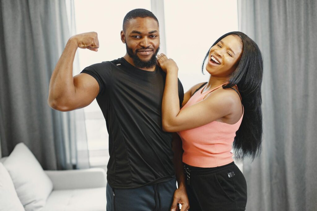 Joyful couple indoors with man flexing bicep and woman laughing, conveying happiness and strength.