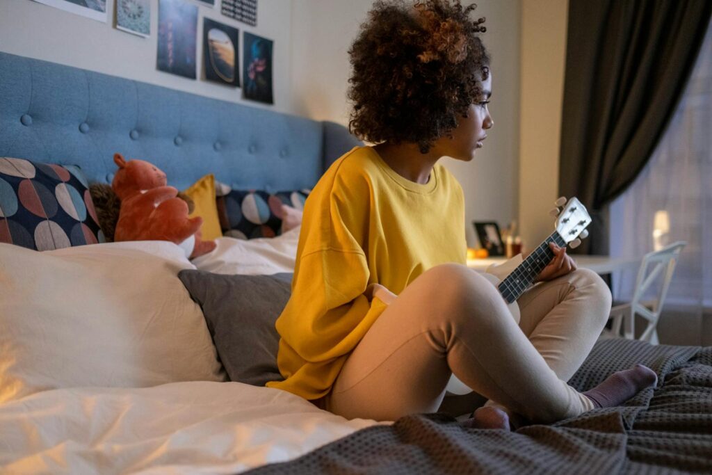 Young girl playing ukulele while sitting on a bed in a cozy, warmly lit bedroom.