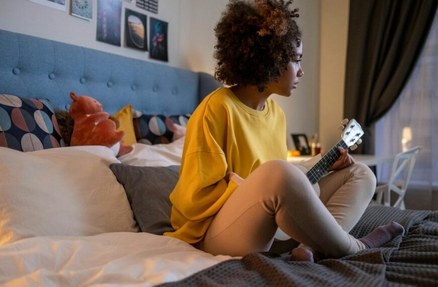 Young girl playing ukulele while sitting on a bed in a cozy, warmly lit bedroom.