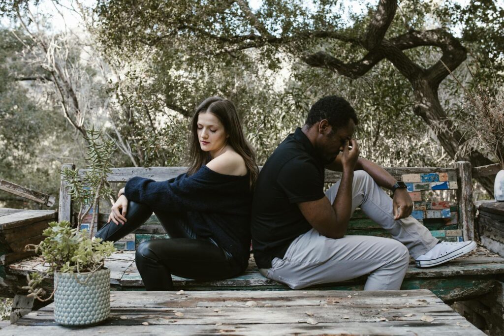 A thoughtful couple sits back-to-back on a rustic bench surrounded by nature, conveying emotions.
