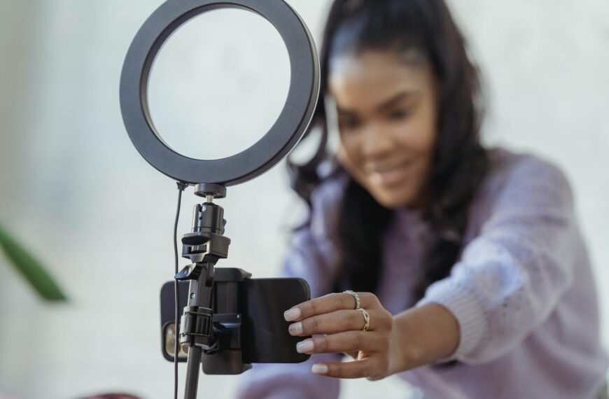 Cheerful young African American female blogger in stylish sweater smiling while setting up camera of smartphone attached to tripod with ring light before recording vlog