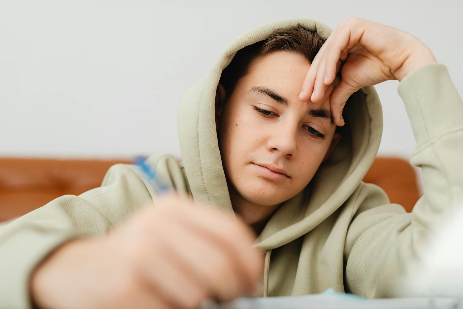 Teenage boy in hoodie writing homework at home, appearing thoughtful and focused.