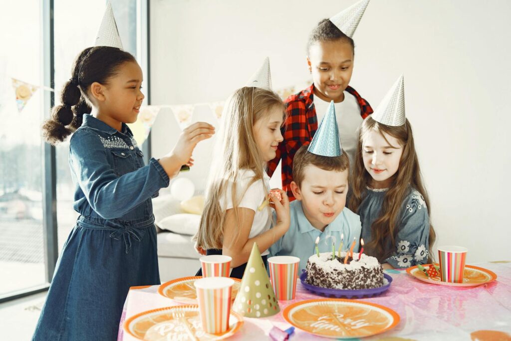 A joyful group of kids celebrating a birthday indoors with cake and party hats.