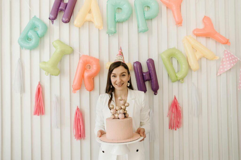 Woman celebrating birthday with colorful balloon decorations and a cake indoors.