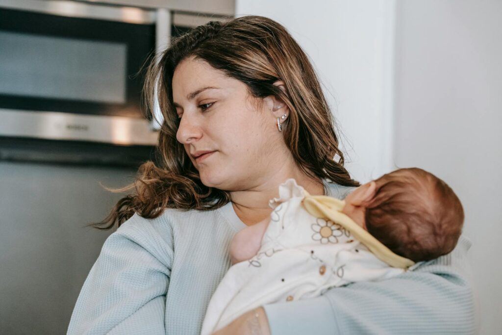 Caring mother with curly hair and cute infant baby in arms standing in light kitchen with microwave oven at home