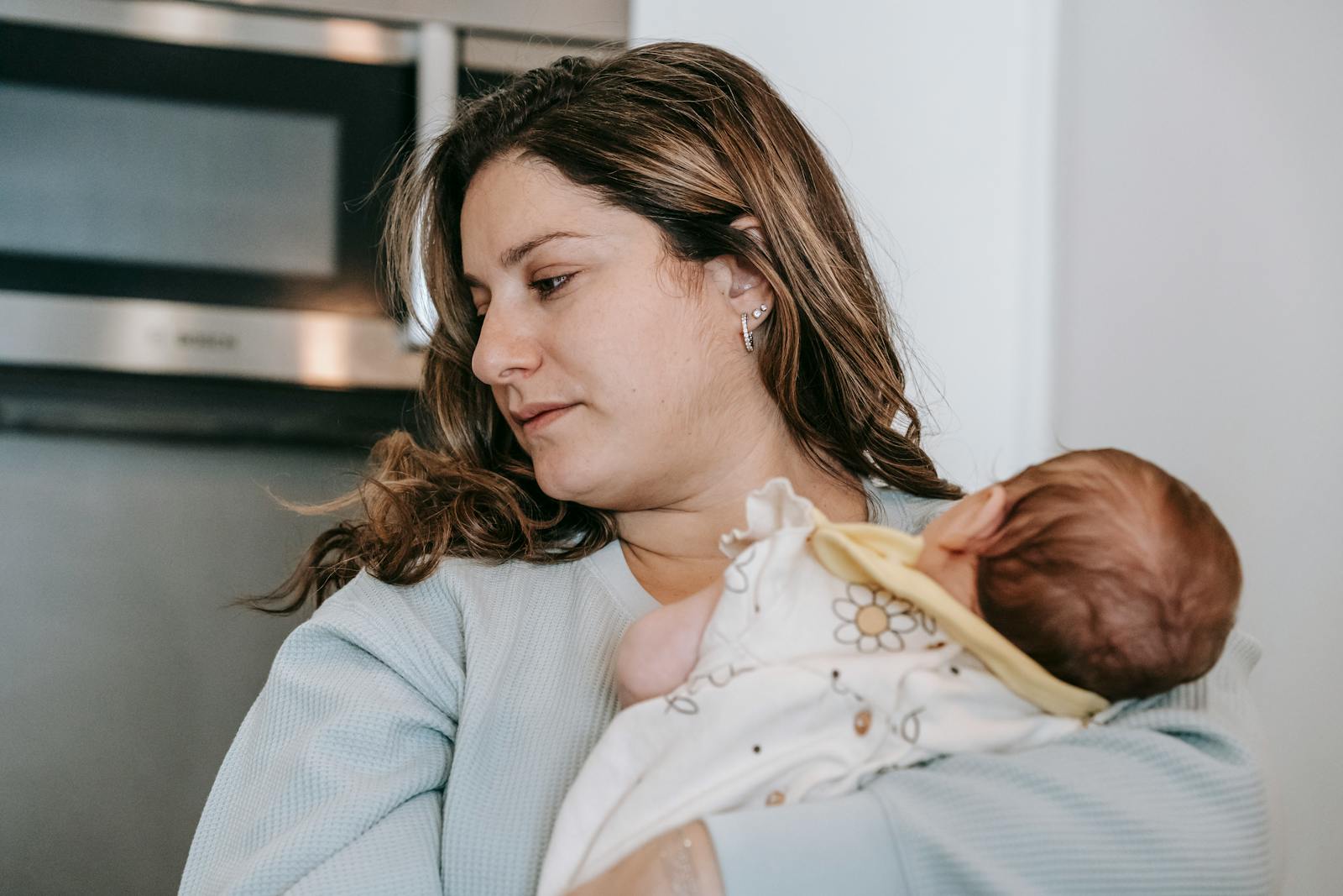 Caring mother with curly hair and cute infant baby in arms standing in light kitchen with microwave oven at home