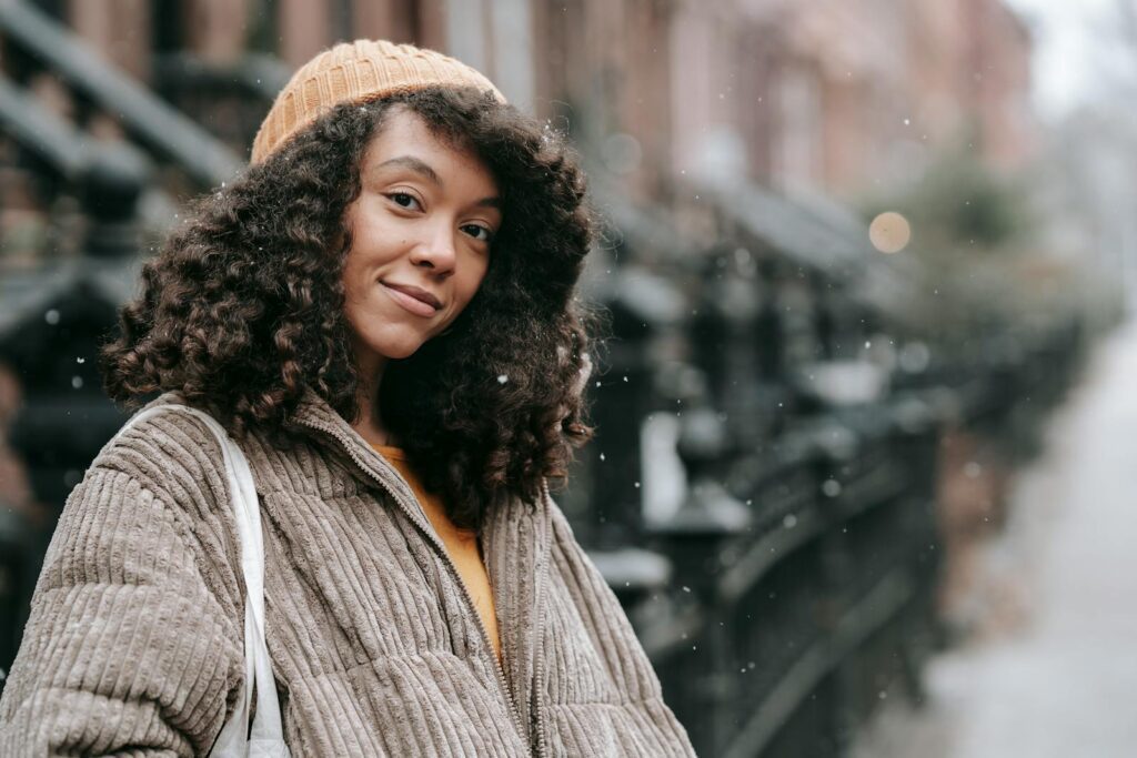 Sincere young ethnic female with Afro hairstyle in warm clothes looking at camera in snowfall in city
