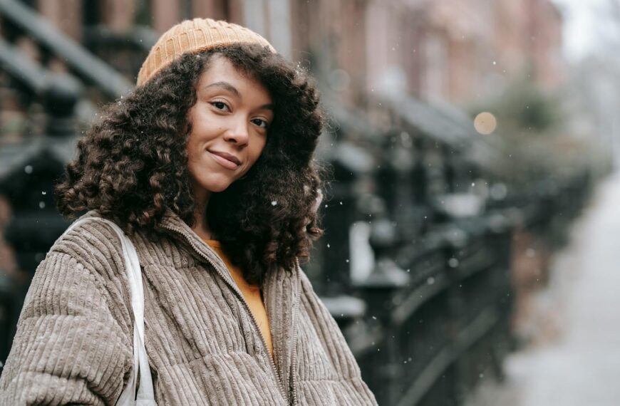 Sincere young ethnic female with Afro hairstyle in warm clothes looking at camera in snowfall in city