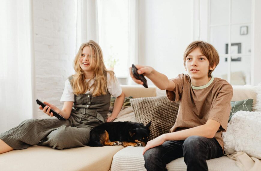 Two kids sitting on a couch with their pet dog, watching TV indoors.