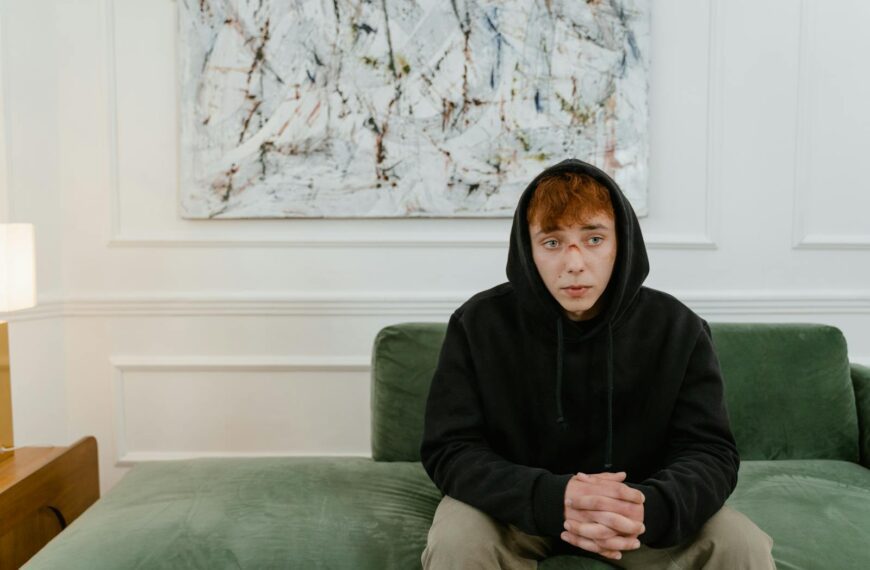 A young man in a black hoodie sits thoughtfully on a green couch indoors.