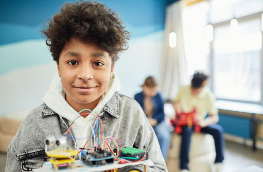 A young boy smiling confidently while holding a DIY robotics project in a classroom setting.