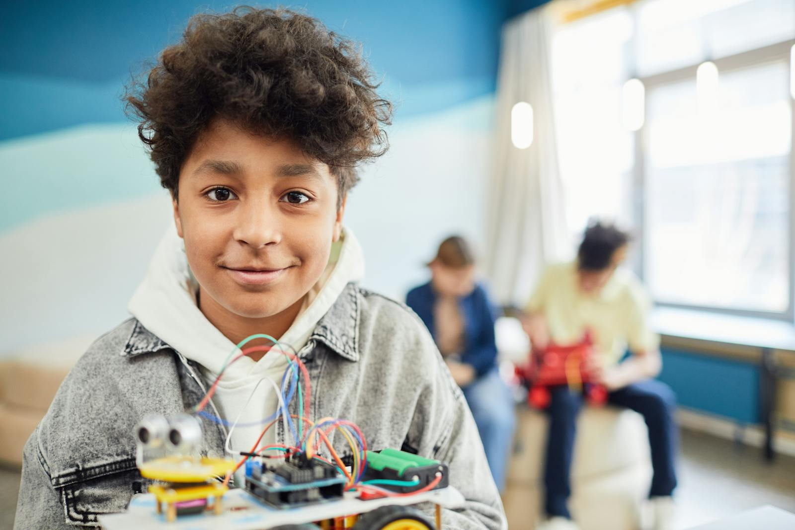 A young boy smiling confidently while holding a DIY robotics project in a classroom setting.