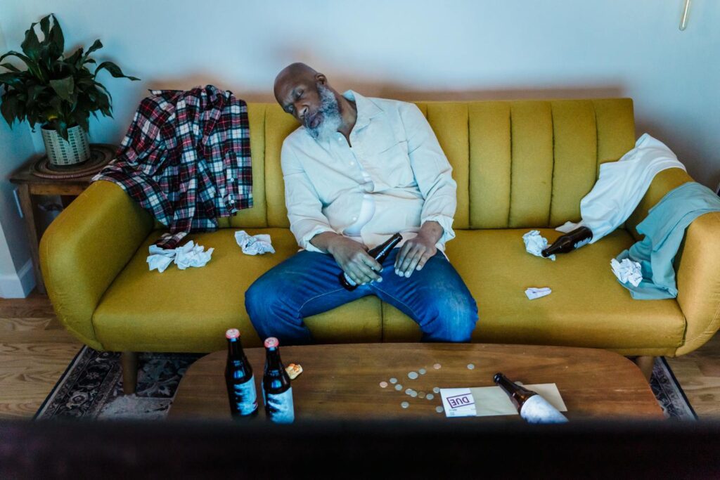An adult man sleeping on a sofa surrounded by beer bottles and crumpled papers indoors.