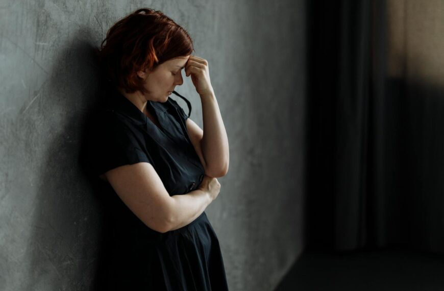 A woman in stress leaning against a concrete wall with eyes closed, conveying deep emotion.