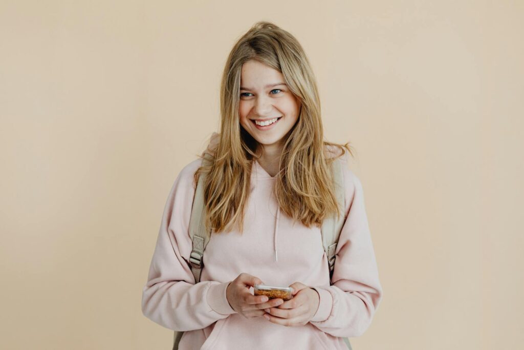 A cheerful teenager with blonde hair wearing a pink sweater and using a smartphone indoors.